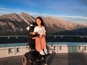 A photo of Tori sitting in her wheelchair on the observation deck at Sulphur Mountain. The mountains are towering over her in the background