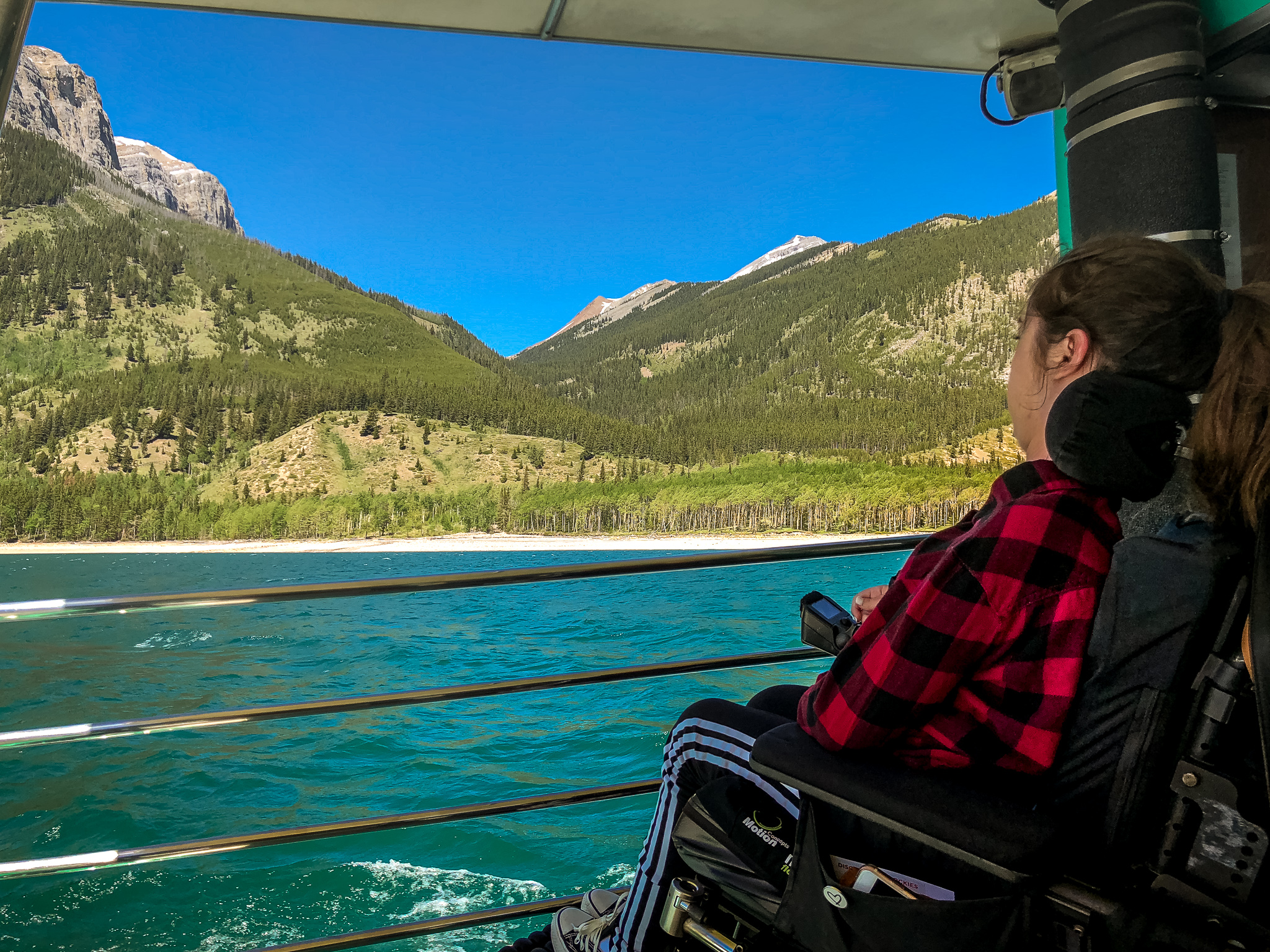 Tori sitting in her wheelchair on the Lake Minnewanka boat cruise. The water is a bright turquoise blue and there are luscious green mountains in the background