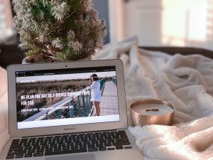A laptop is placed on top of a fluffy white blanket. On the screen of the laptop is the homepage to Guess Where Trips website. To the right of the laptop is a gold candle. In the background, there is a miniature Christmas tree.