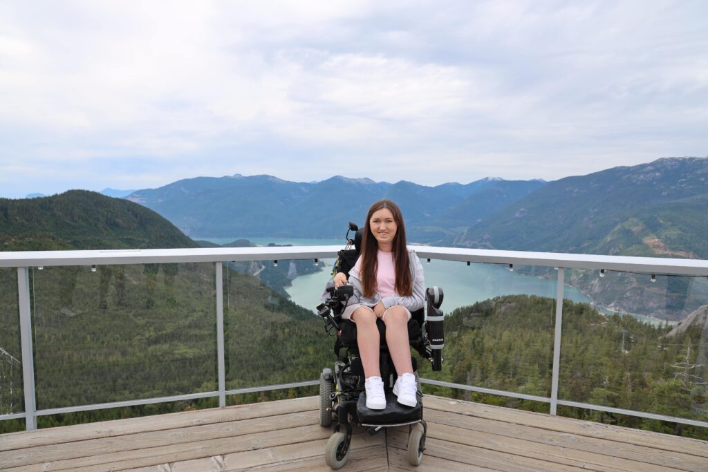 Tori in her wheelchair, looking at the camera and smiling on the viewing platform at the sea to sky gondola. Behind her you can see beautiful scenery of luscious forests, water and mountains