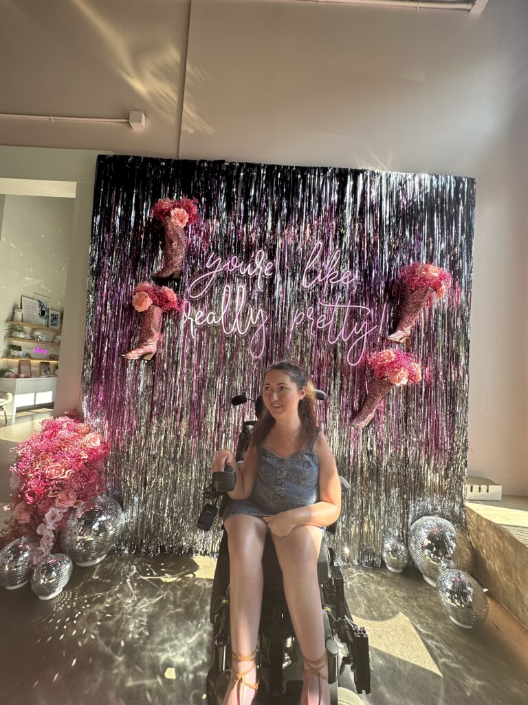 Tori sitting in her wheelchair in front of an aesthetic photo backdrop that says "you're like really pretty” in pink neon lights. There are several large disco balls on the floor.