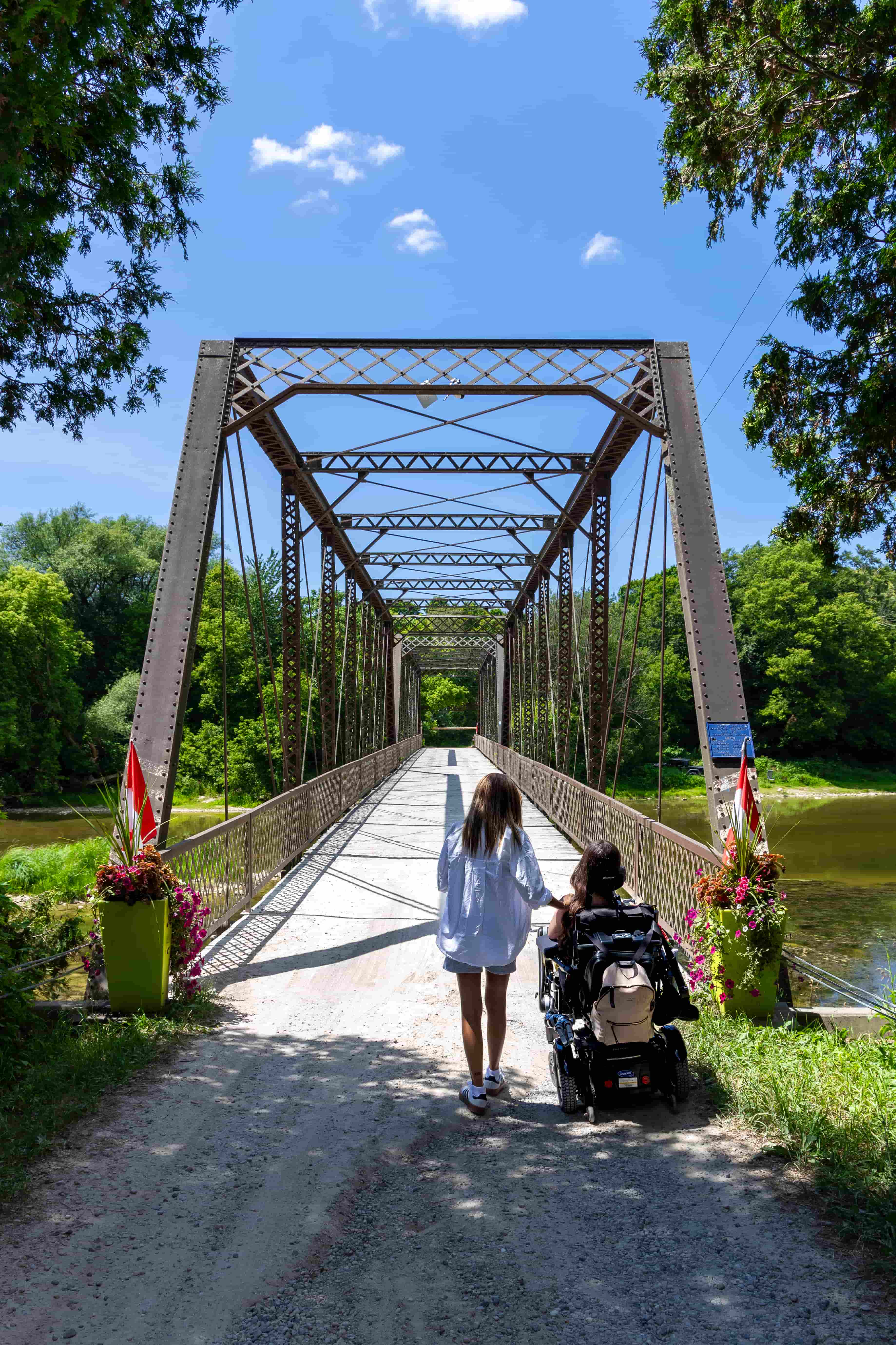 Tori, a wheelchair accessible travel blogger, in her power wheelchair alongside her assistant as they stroll across Balls Bridge in Central Huron, Ontario. The bridge is a relaxing, accessible spot to enjoy together.