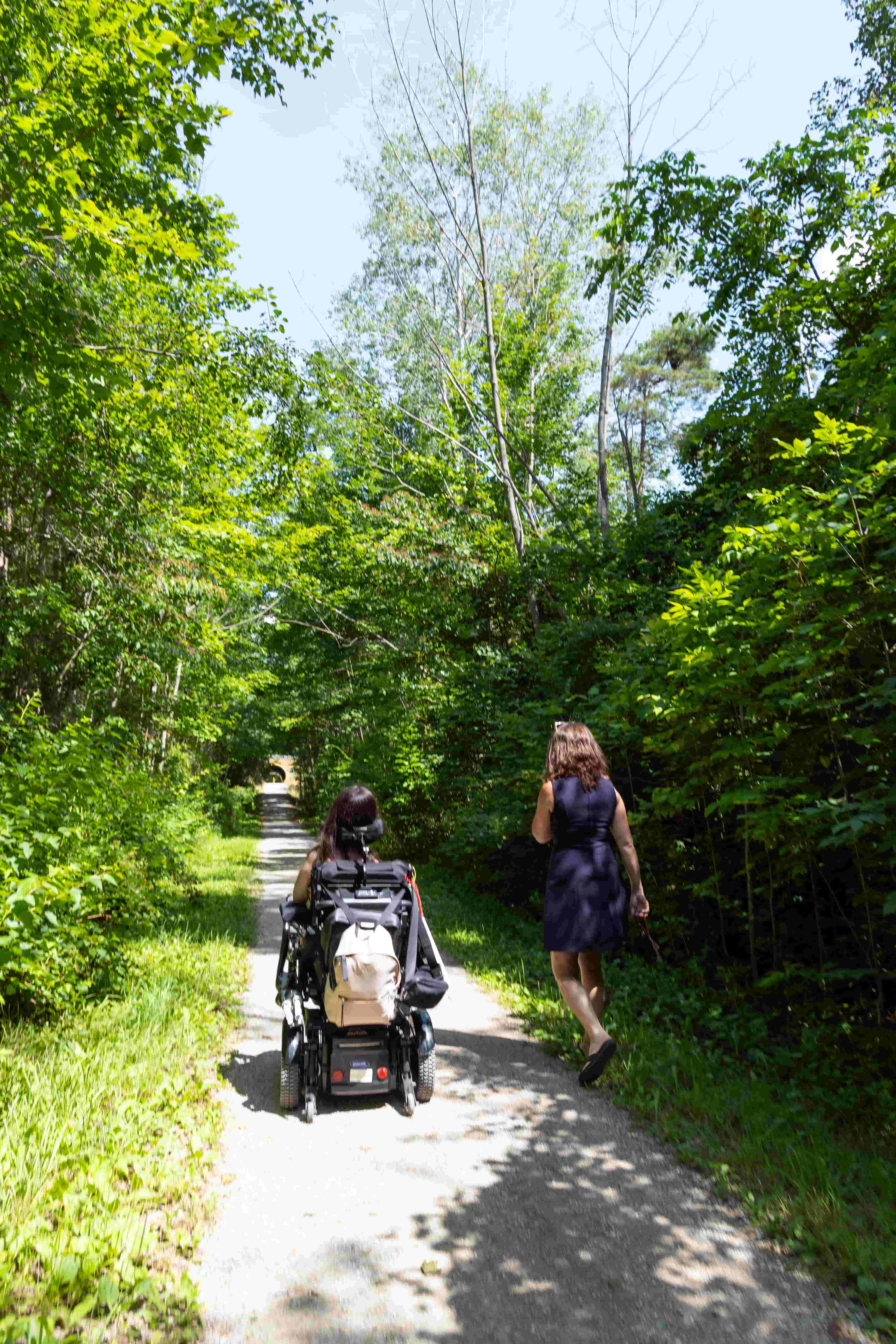 Tori, a wheelchair accessible travel blogger, taking a stroll in her power wheelchair alongside another person on the G2G Rail Trail in Central Huron, Ontario. The trail offers a scenic and accessible outdoor experience.