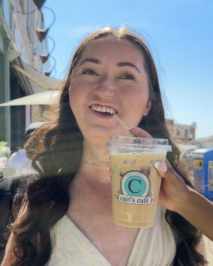 Tori, a wheelchair accessible travel blogger, smiling while her assistant is holding a drink at Cait’s Café in Central Huron, Ontario. The café patio provides a welcoming and accessible spot to relax.
