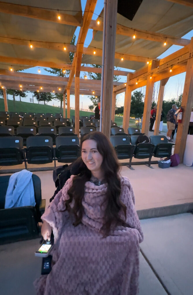 Tori, a wheelchair accessible travel blogger, sitting at the open-air theater in Central Huron, Ontario, with a pink blanket wrapped around her and a smile on her face. The venue offers an accessible and cozy way to enjoy live performances.