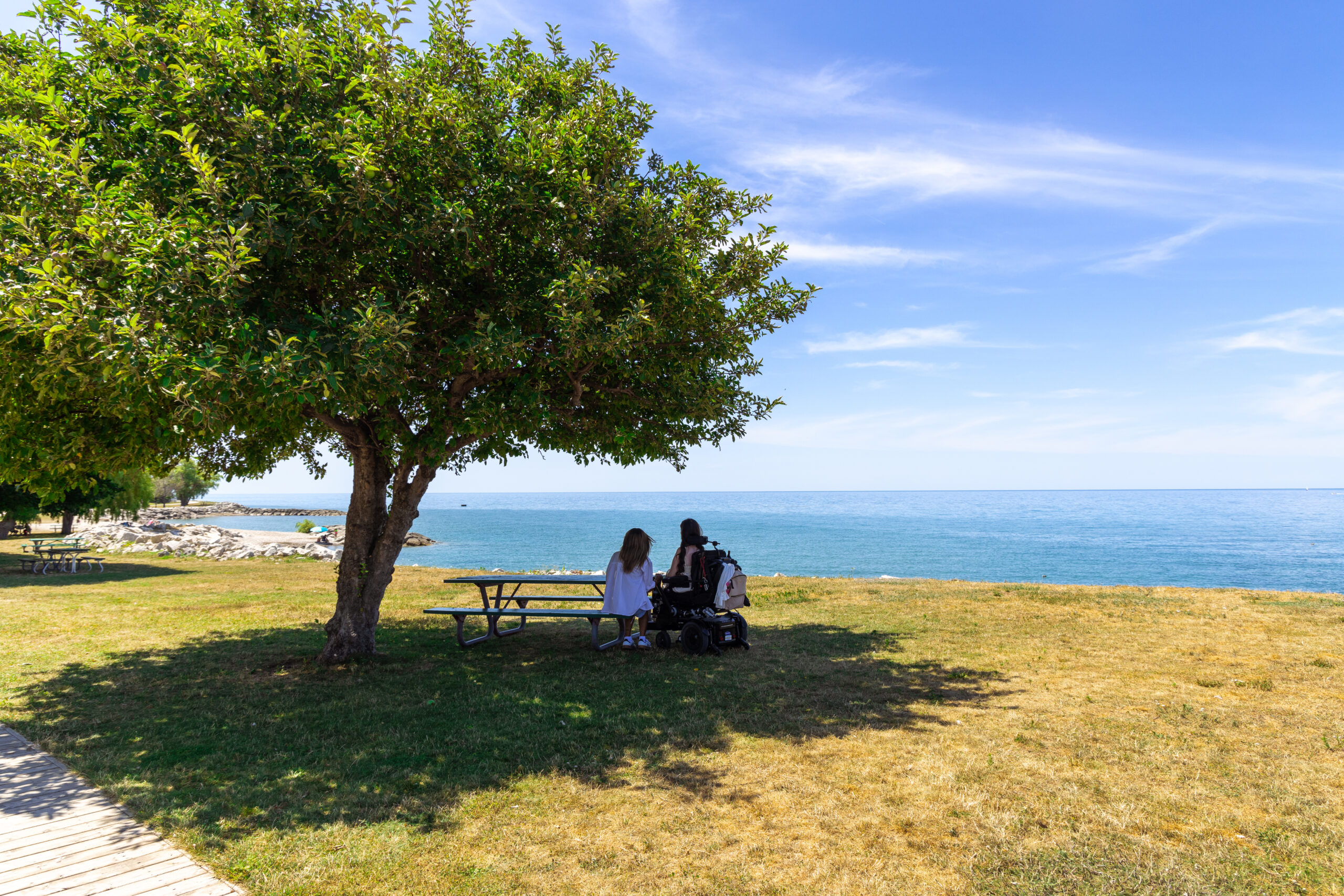 Tori, a wheelchair accessible travel blogger, sitting with her assistant at a picnic table overlooking the lake at a Central Huron beach. The photo captures the beauty of Ontario’s waterfront and highlights accessible travel experiences.
