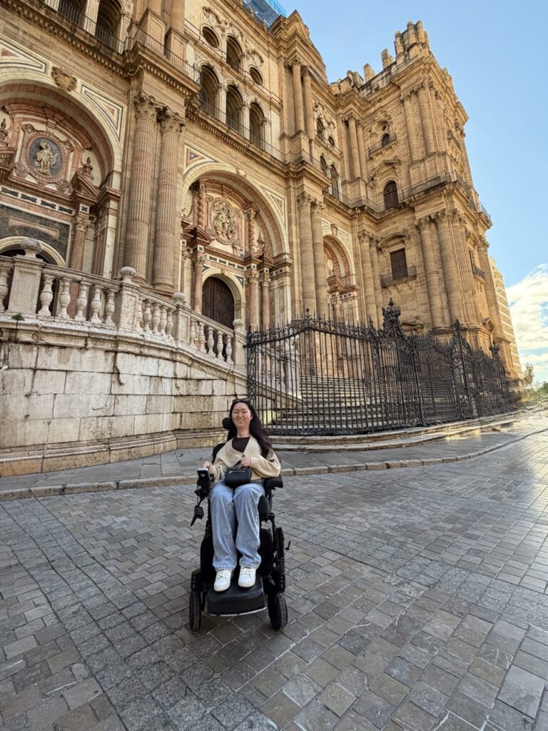 Tori Hunter, a wheelchair user and disabled travel blogger, poses in her power wheelchair in front of the ornate Baroque facade of Málaga Cathedral in Spain