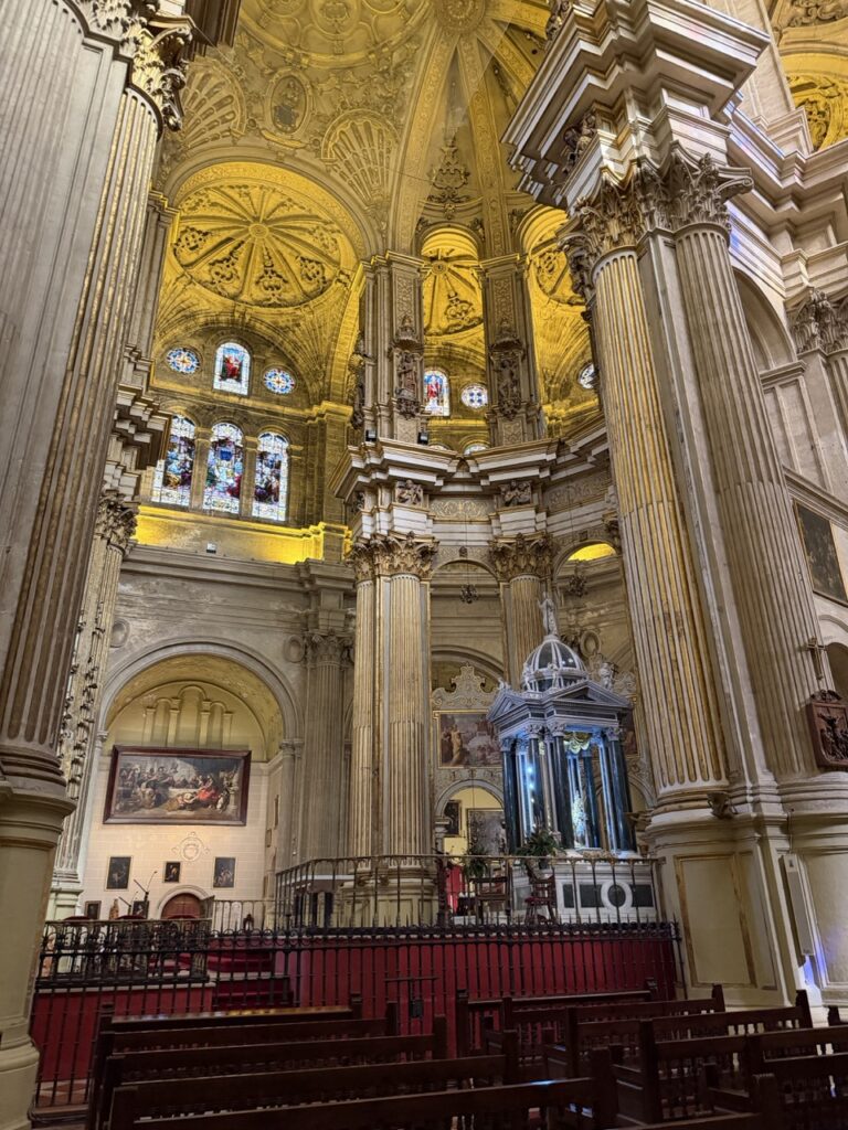 The grand wheelchair accessible interior of Málaga Cathedral in Malaga, Spain, featuring towering stone columns, golden vaulted ceilings, stained glass windows, and an elaborate main altar