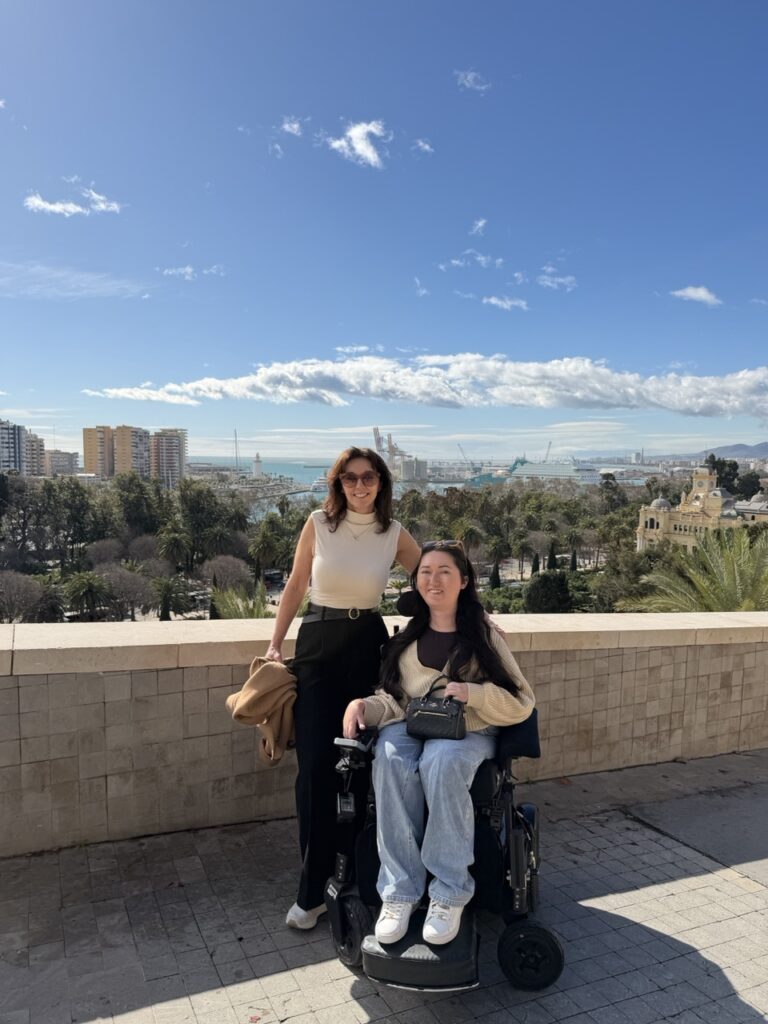 Tori Hunter, a wheelchair accessible travel blogger, posing in her power wheelchair with a companion on the accessible terrace of Alcazaba fortress in Malaga, Spain, with panoramic views of the city, port, and Mediterranean Sea behind them