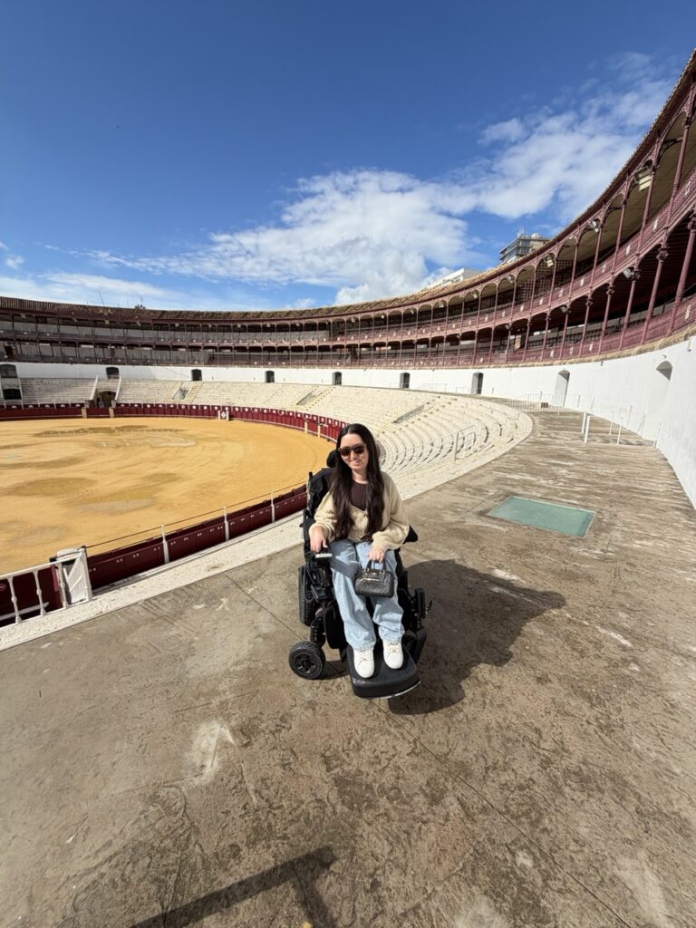 Tori Hunter, a wheelchair accessible travel blogger, sitting in her power wheelchair on the arena floor of the Plaza de Toros bullring in Malaga, Spain, a wheelchair accessible attraction on the Costa del Sol