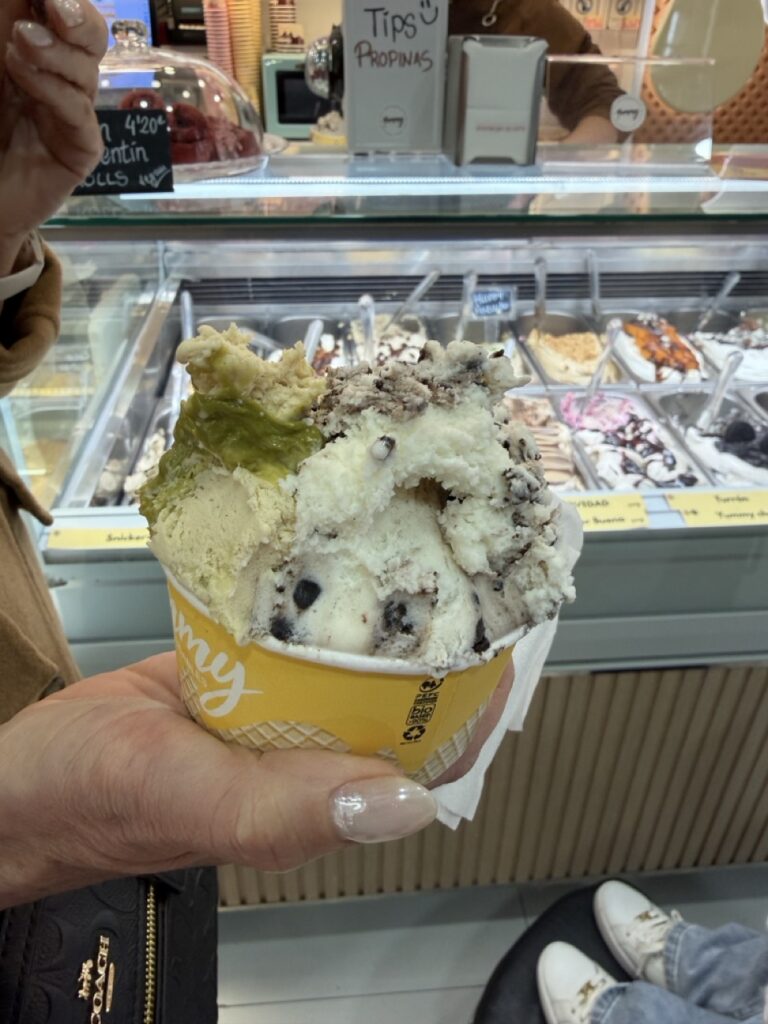 A cup of gelato with pistachio and cookies and cream flavours held in front of the gelato display counter at Yummy Helados Saludables, a wheelchair accessible ice cream shop in Malaga, Spain
