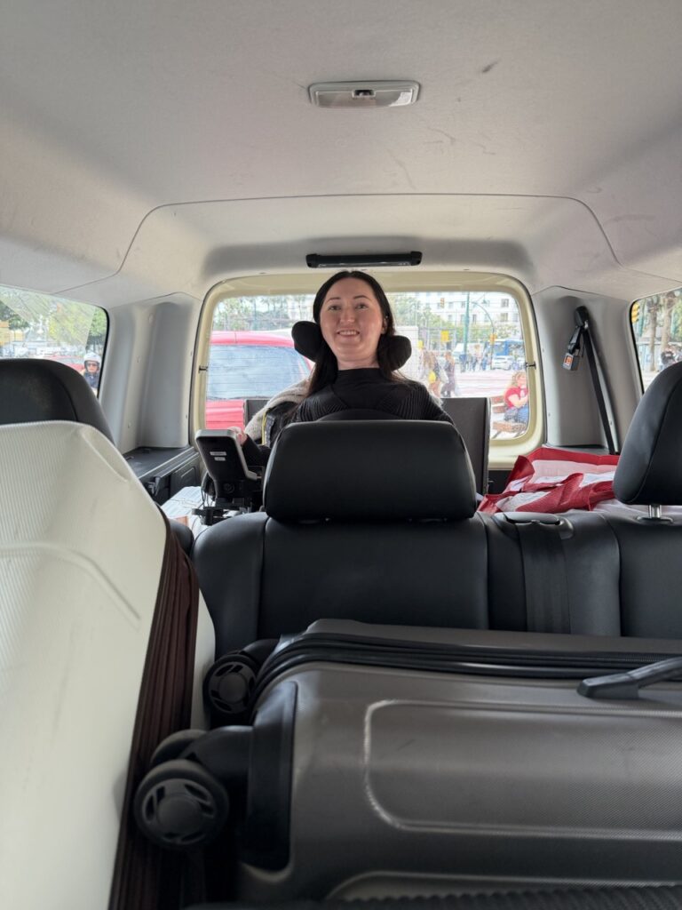 Tori Hunter, a wheelchair accessible travel blogger, seated in her power wheelchair inside a wheelchair accessible taxi van in Malaga, Spain, smiling with luggage visible in the foreground
