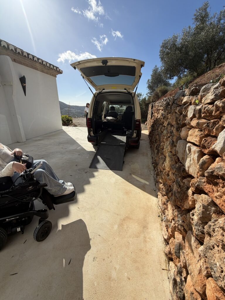 A wheelchair accessible van with a rear entry ramp deployed on a sunny day in the Malaga region of Spain, with a power wheelchair user visible in the foreground preparing to board