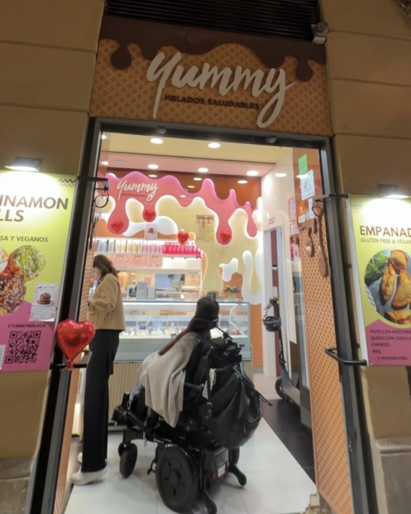 Tori Hunter, a wheelchair accessible travel blogger, entering Yummy Helados Saludables in her power wheelchair through a step-free entrance — a wheelchair accessible ice cream shop in Malaga, Spain