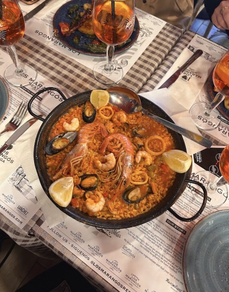A large pan of seafood paella with prawns, mussels, squid, and lemon served at Los Marangós tapas restaurant in Malaga, Spain, with Aperol Spritz glasses on the table