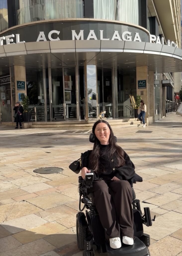 Tori Hunter, a wheelchair accessible travel blogger, smiling in her power wheelchair outside the entrance of AC Hotel Malaga Palacio by Marriott — a wheelchair accessible hotel in Malaga, Spain
