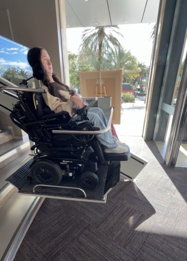 Tori Hunter using a platform lift in her power wheelchair inside the lobby of AC Hotel Malaga Palacio in Malaga, Spain — an example of the wheelchair accessibility features available at the hotel