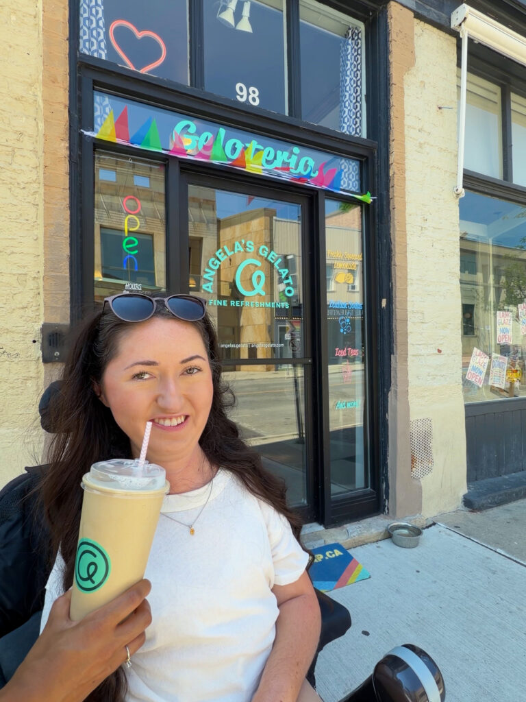 Tori Hunter, a wheelchair user and disabled travel blogger, smiling and holding an iced drink outside Angela's Gelato, a wheelchair accessible café in downtown Stratford, Ontario