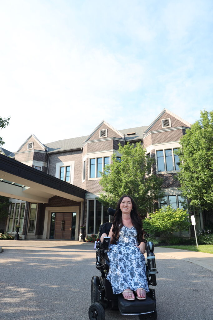  Tori Hunter, a wheelchair user and disabled travel blogger, smiling in her power wheelchair outside The Bruce Hotel, a wheelchair accessible boutique hotel in Stratford, Ontario
