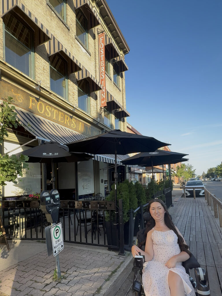 Tori Hunter, a wheelchair user and disabled travel blogger, smiling in her power wheelchair on the accessible boardwalk outside Foster's Inn restaurant in downtown Stratford, Ontario