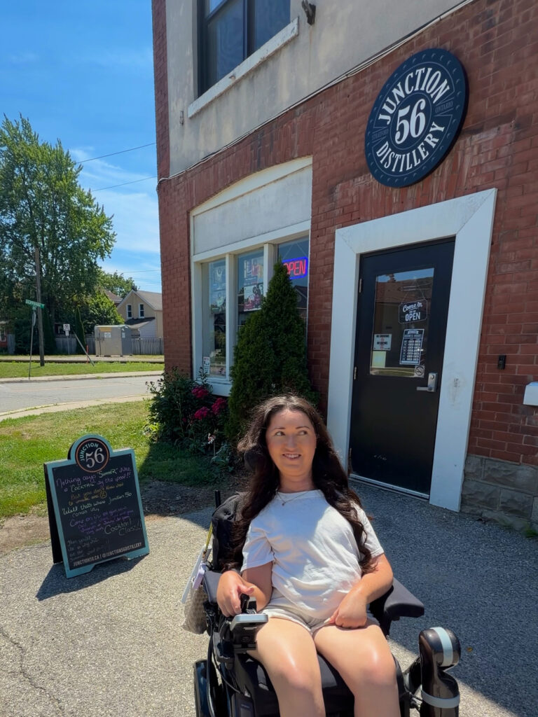 Tori Hunter, a wheelchair user and disabled travel blogger, smiling in her power wheelchair outside the entrance of Junction 56 Distillery, a wheelchair accessible distillery in Stratford, Ontario