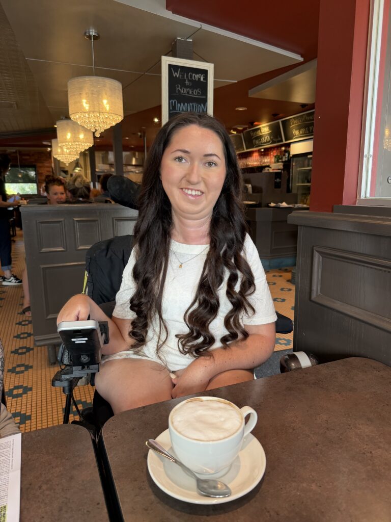 Tori Hunter, a wheelchair user and disabled travel blogger, smiling in her power wheelchair at a table with a cappuccino inside Romeo's, a wheelchair accessible restaurant in Stratford, Ontario