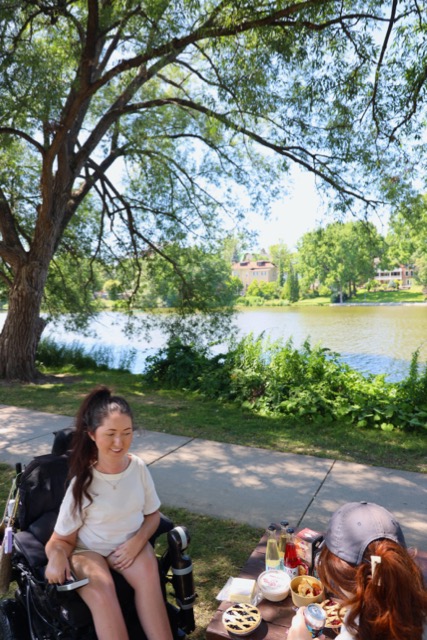 Tori Hunter, a wheelchair user and disabled travel blogger, enjoying an accessible riverside picnic with local pies and cheese along the Avon River near Shakespearean Gardens in Stratford, Ontario