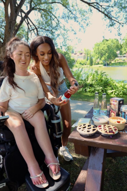 Tori Hunter, a wheelchair user and disabled travel blogger, laughing with a friend in her power wheelchair at an accessible picnic table along the Avon River in Stratford, Ontario