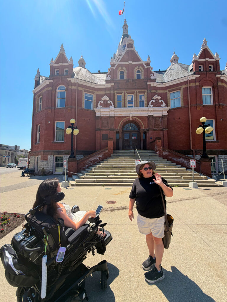 Tori Hunter, a wheelchair user and disabled travel blogger, meeting her tour guide in her power wheelchair in front of Stratford City Hall, a historic landmark in downtown Stratford, Ontario