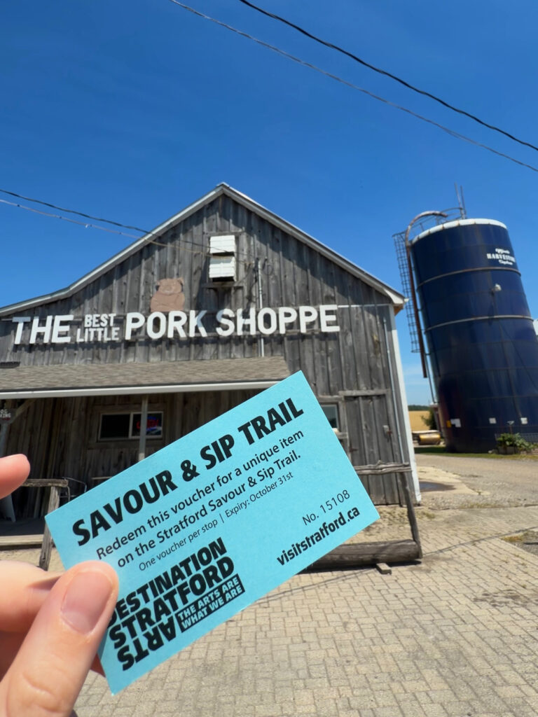 Tori Hunter, a wheelchair user and disabled travel blogger, holding a Savour & Sip Trail voucher in her power wheelchair outside The Best Little Pork Shoppe on the Stratford Savour & Sip Trail in Stratford, Ontario