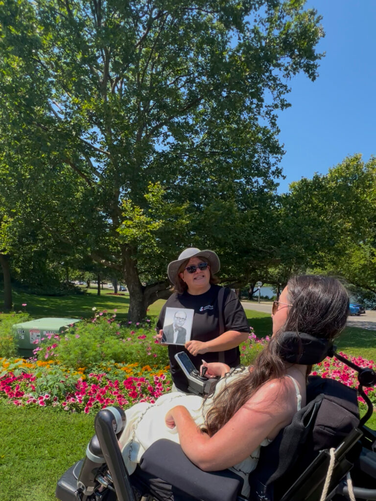 Tori Hunter, a wheelchair user and disabled travel blogger, taking part in an accessible walking tour in Stratford, Ontario, as a local guide shares a historical photo near the gardens along the Avon River