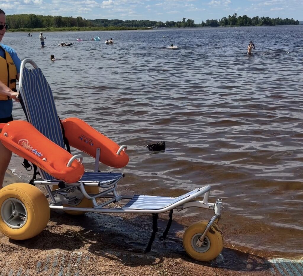 Beach wheelchair with balloon tires and floating pontoons at Petawawa Point Beach in Petawawa, Ontario, with swimmers and the lake shoreline in the background — an example of accessible beach access for wheelchair users in Ontario and Canada