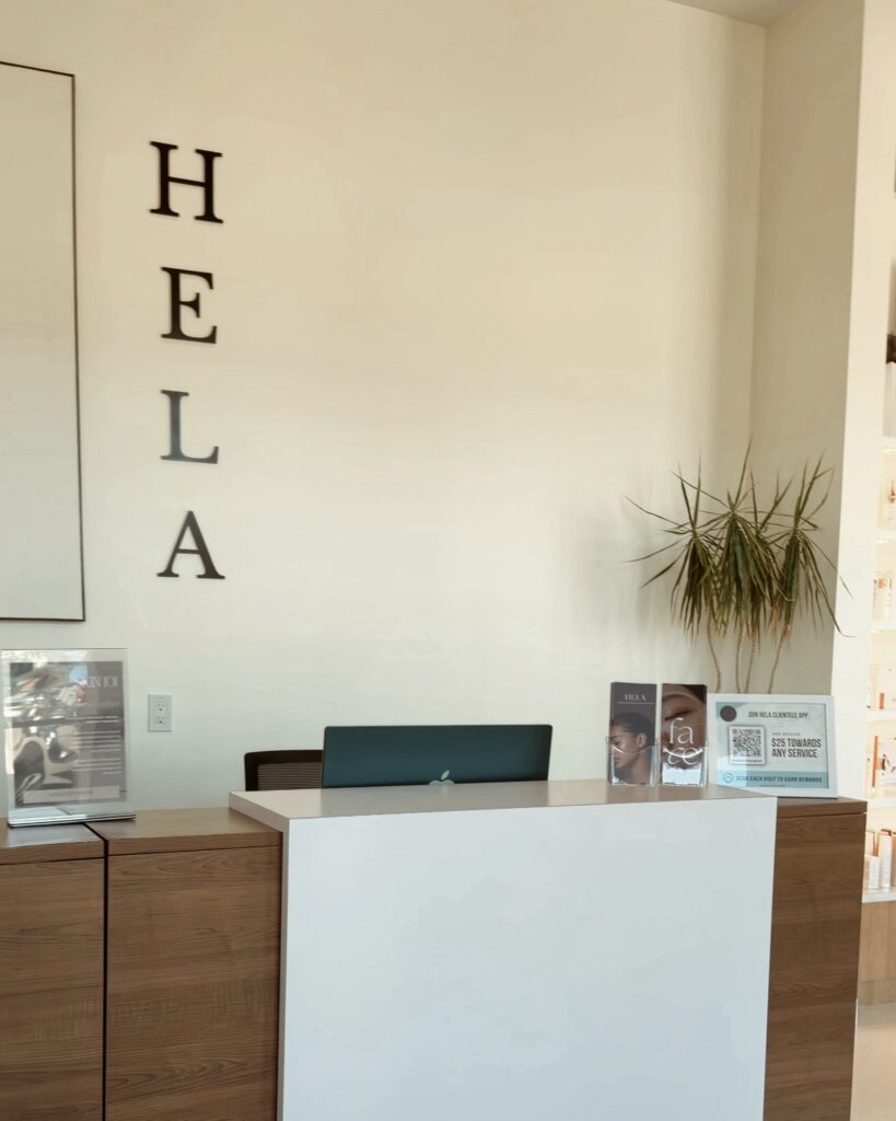 Reception desk at HELA skin studio with wall-mounted "HELA" signage, wood-panelled counter at wheelchair-accessible height, and QR code display for the clientele rewards app.