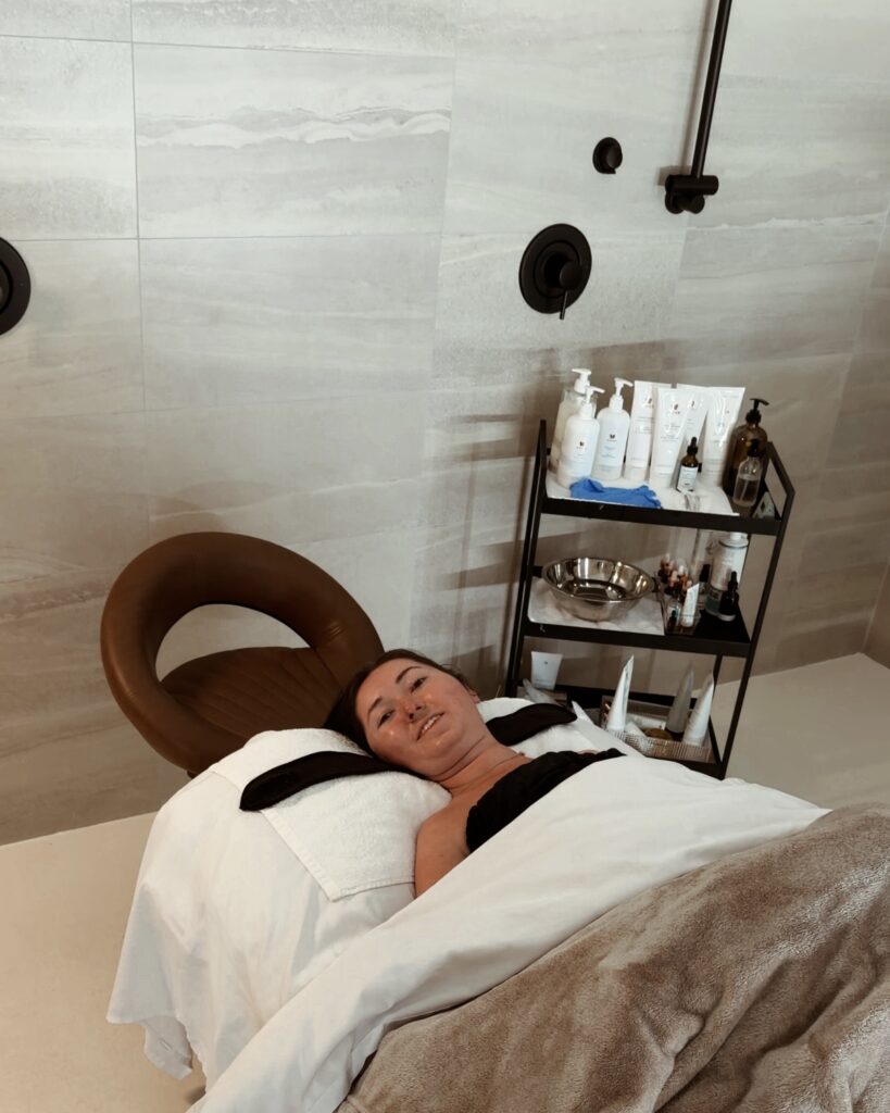 Woman in a power wheelchair reclined for a facial treatment at HELA skin studio, with a rolling cart of skincare products beside the accessible treatment bed.