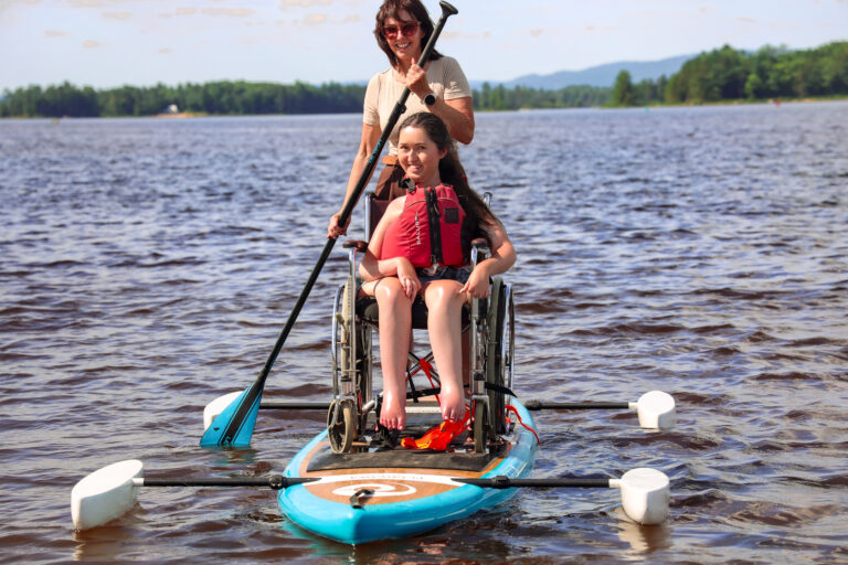 A manual wheelchair secured onto the free adaptive stand-up paddleboard at Petawawa Point Beach in Ontario, with a staff member paddling from behind on the Ottawa River.