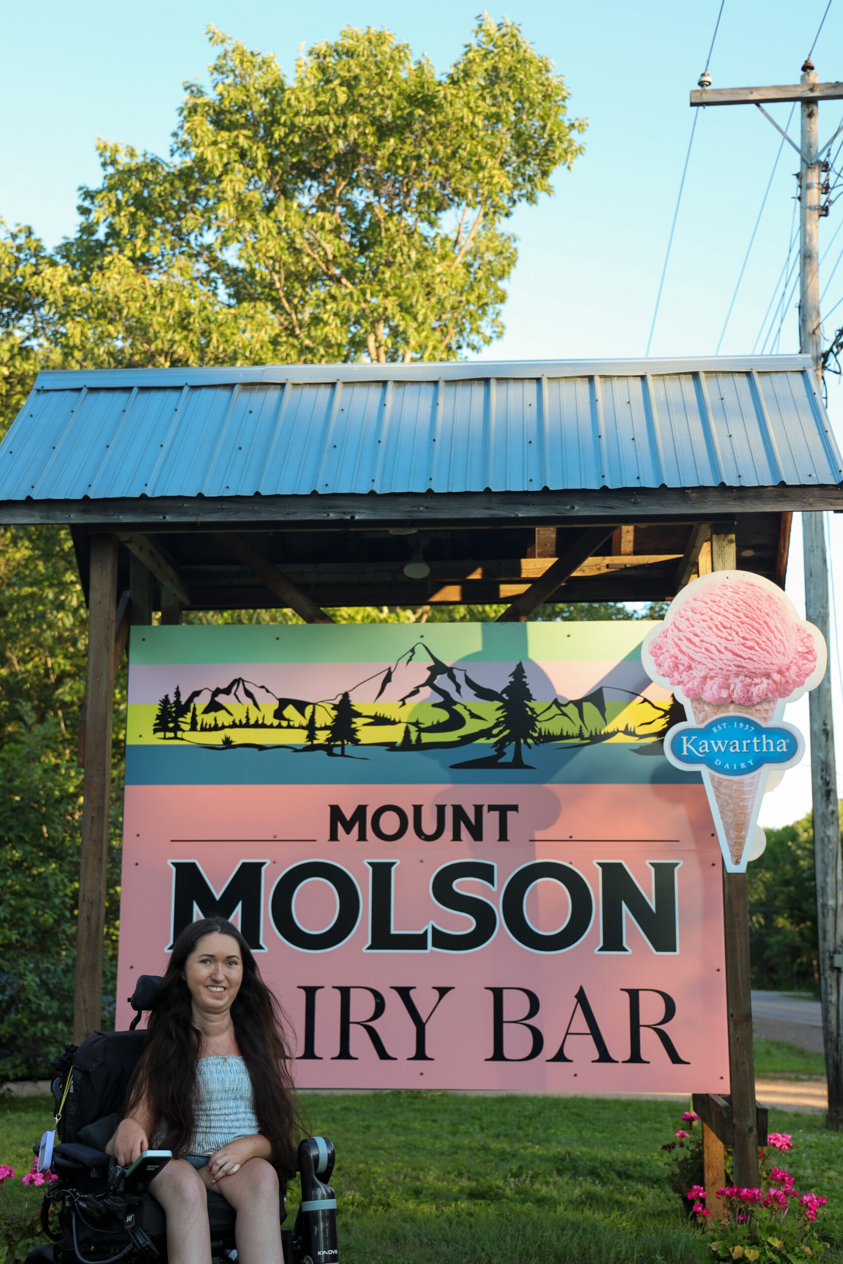 Tori Hunter, a wheelchair user and disabled travel blogger, in her power wheelchair in front of the Mount Molson Dairy Bar and Kawartha Dairy sign at the top of the world's shortest downhill ski run in Petawawa, Ontario.