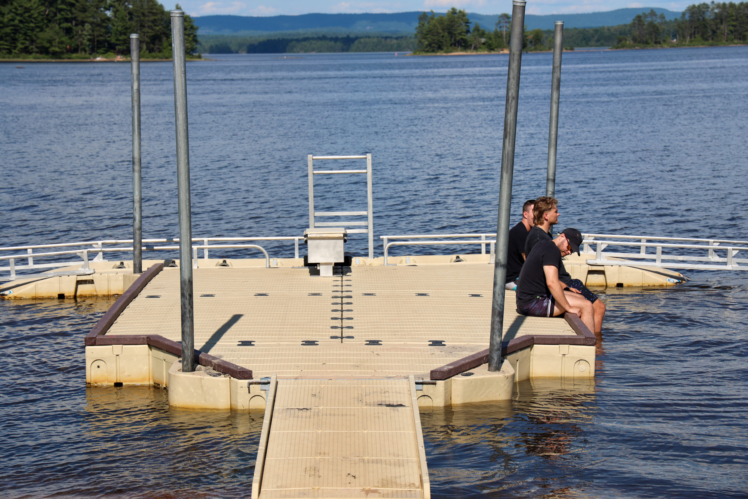 Visitors sitting on a floating kayak dock with their feet in the water on the Ottawa River near Petawawa, Ontario, with forested hills in the background.