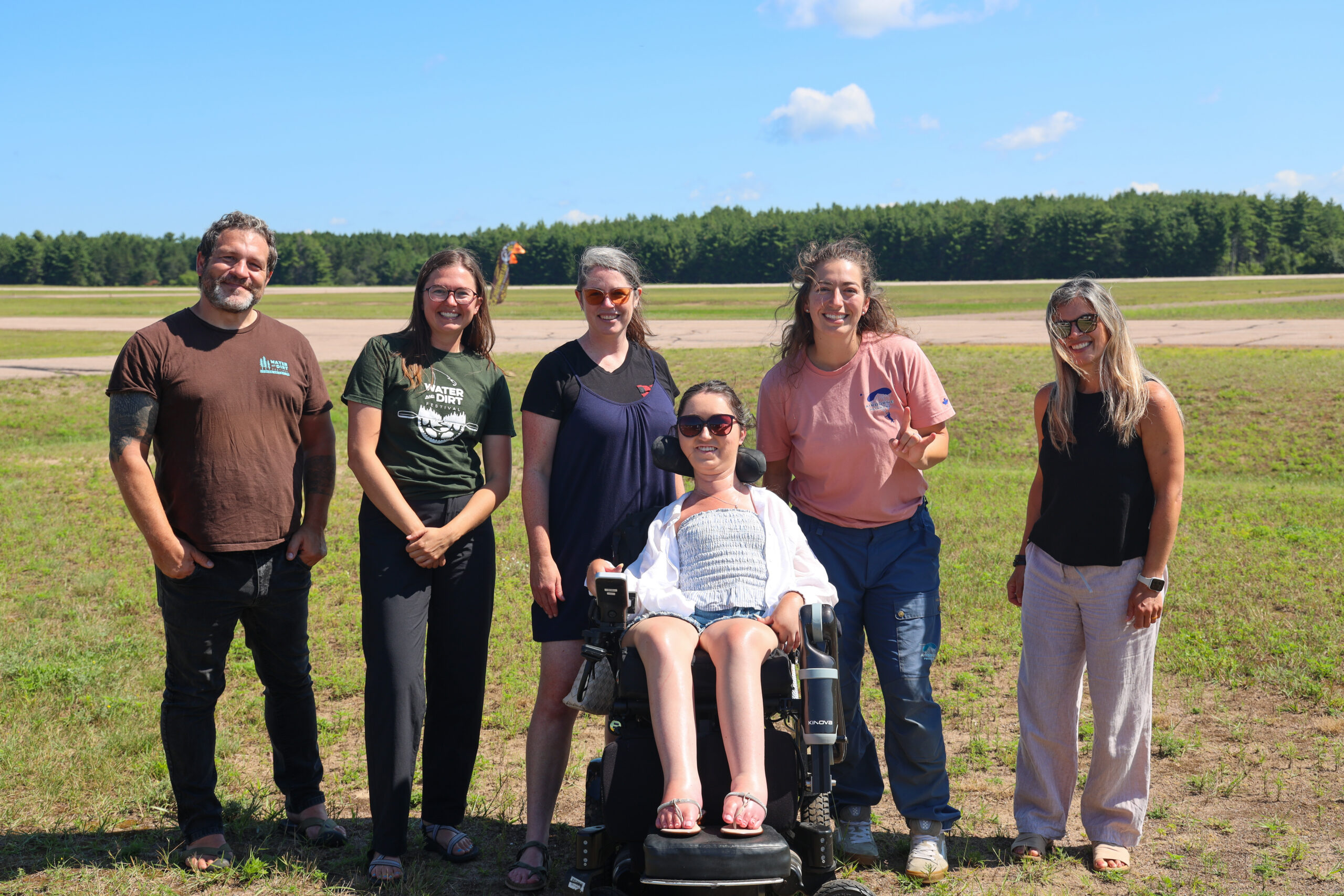 Tori Hunter, a wheelchair user and disabled travel blogger, in her power wheelchair with the Airborne Petawawa team on the grass airfield in Petawawa, Ontario, after learning about their adaptive skydiving harness program.