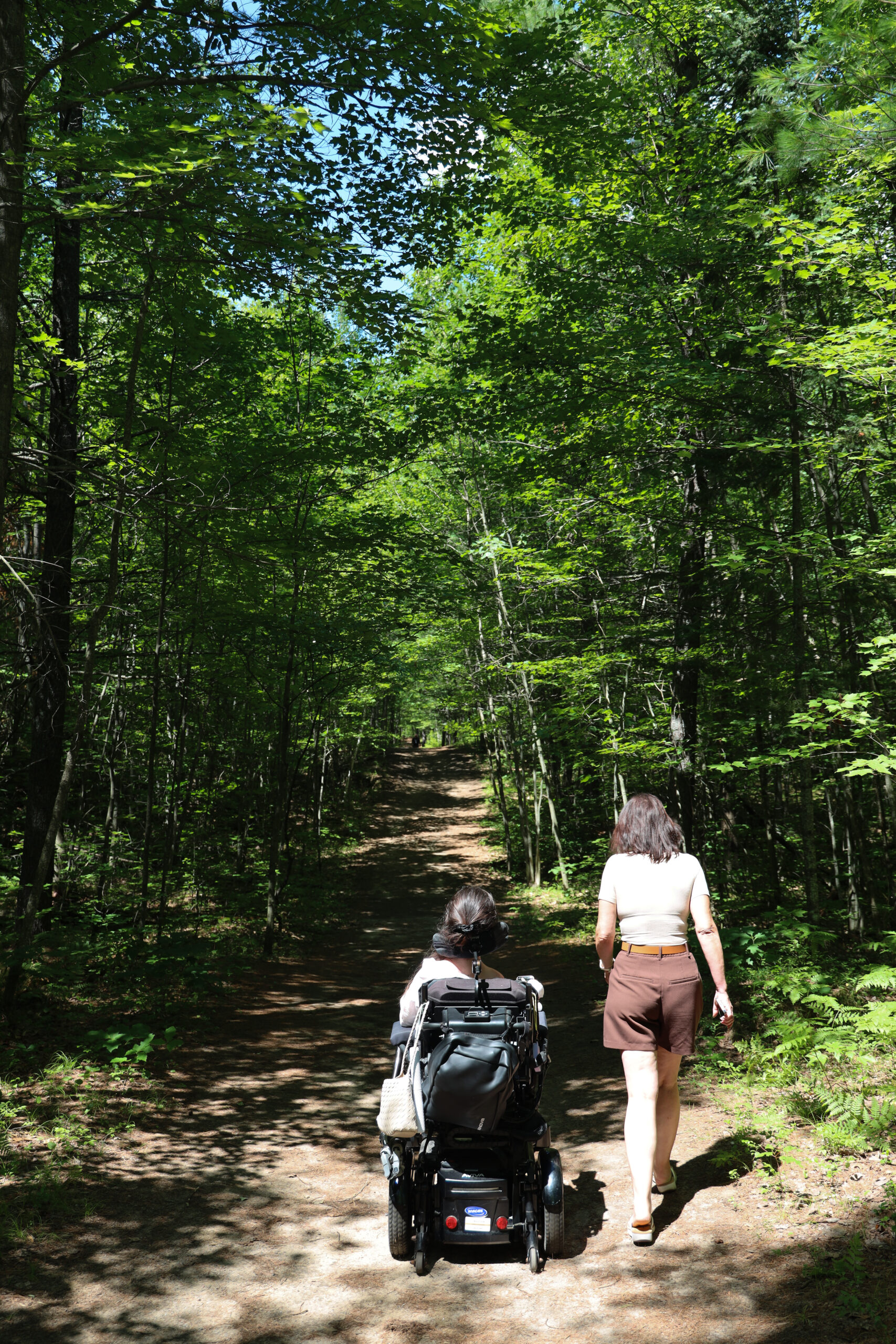 Tori Hunter, a wheelchair user and disabled travel blogger, rolling her power wheelchair alongside a friend walking beside her on the forested Aki Trail at Petawawa Terrace Provincial Park, Ontario.