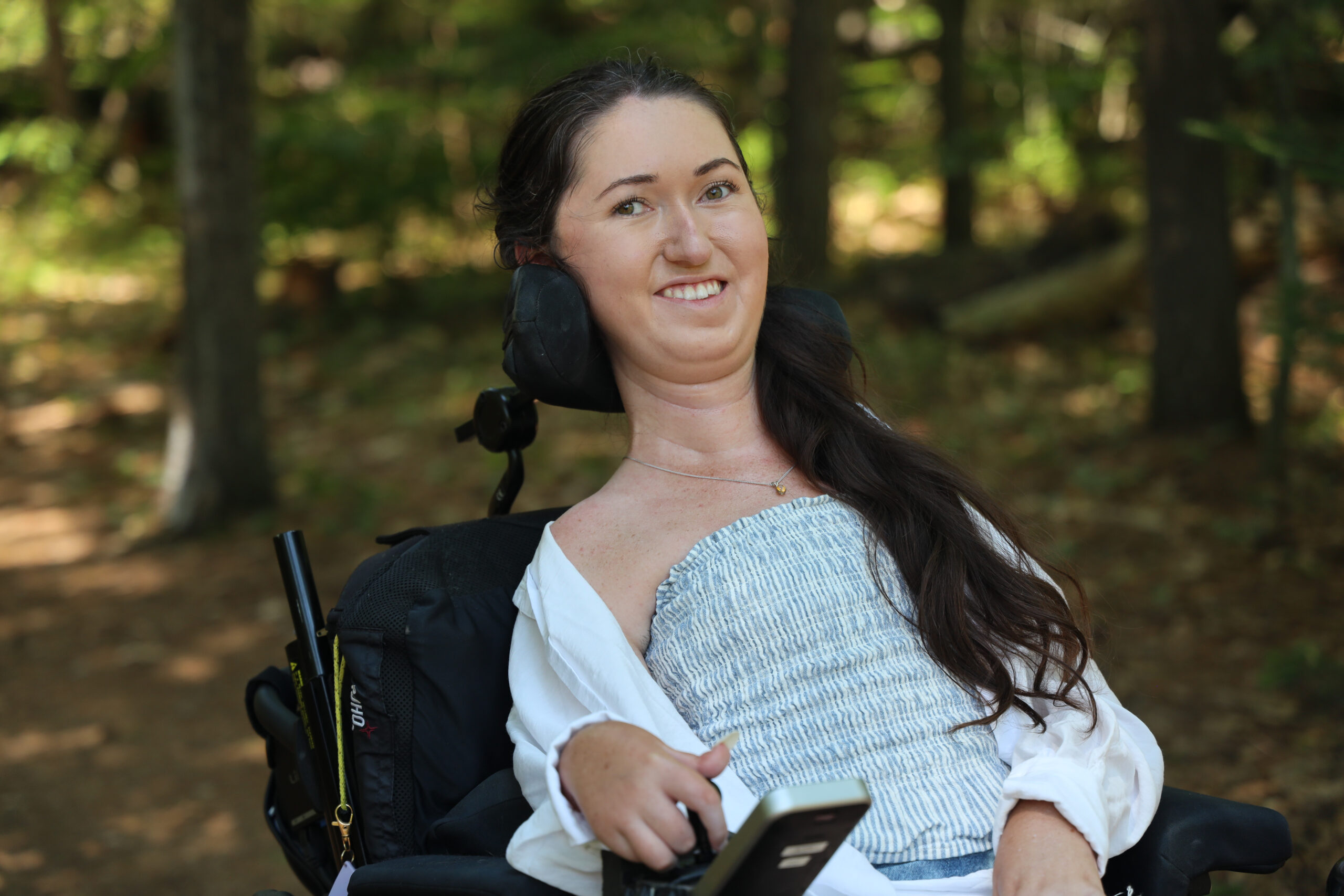 Close-up of Tori Hunter, a wheelchair user and disabled travel blogger, smiling in her power wheelchair along the shaded Aki Trail at Petawawa Terrace Provincial Park in Ontario.