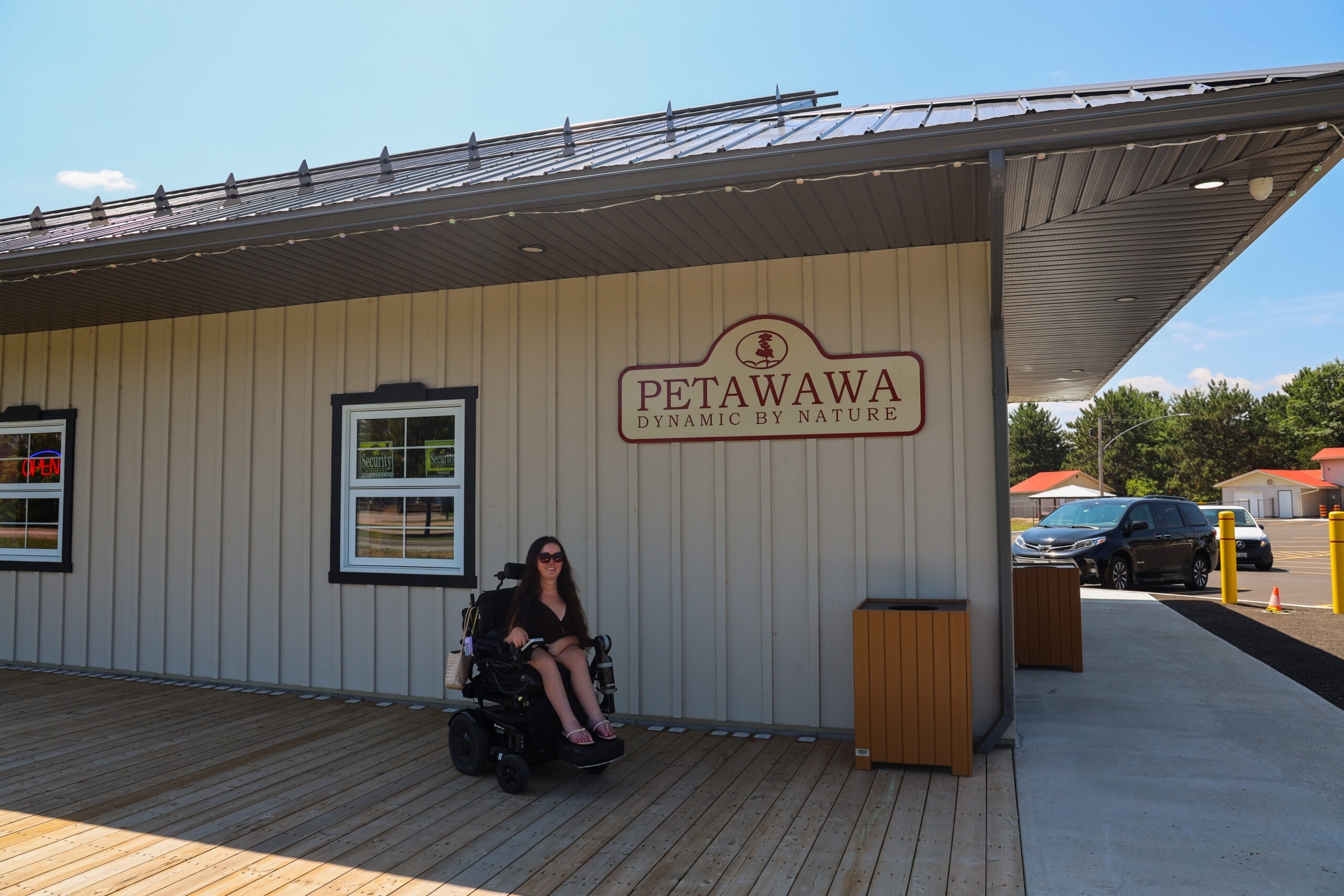 Tori Hunter, a wheelchair user and disabled travel blogger, smiling in her power wheelchair beside the "Petawawa: Dynamic by Nature" sign outside a local shop in Petawawa, Ontario.