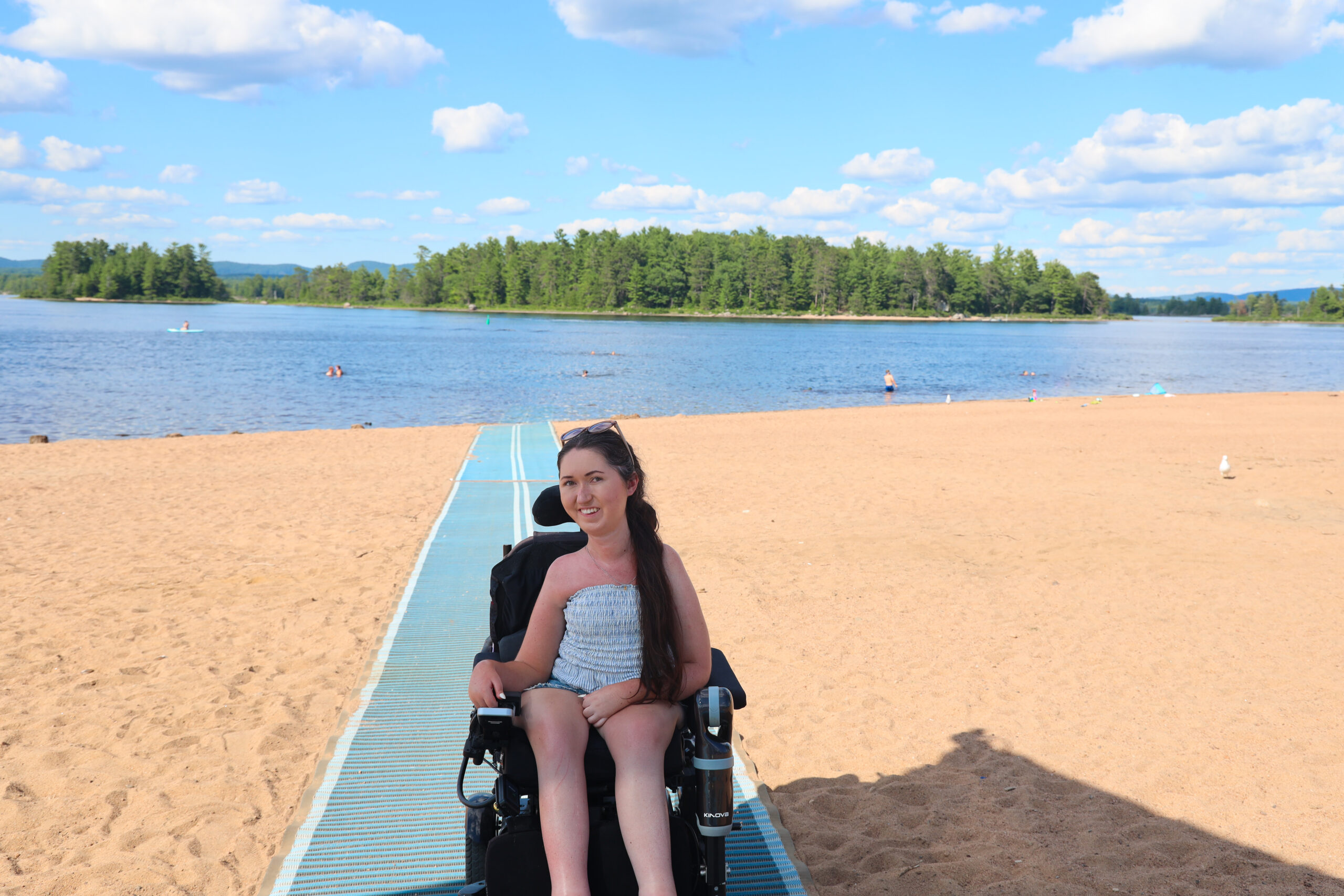 Tori Hunter, a wheelchair user and disabled travel blogger, sitting in her power wheelchair on the blue Mobi-Mat that runs from the sand straight into the water at Petawawa Point Beach, one of the most wheelchair accessible beaches in Ontario.