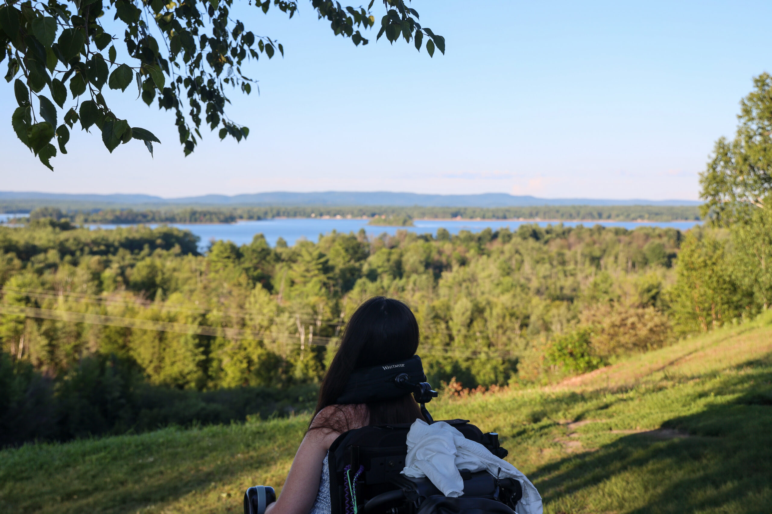Tori Hunter, a wheelchair user and disabled travel blogger, looking out over the Ottawa River from a grassy lookout point near Petawawa, Ontario, in her power wheelchair.