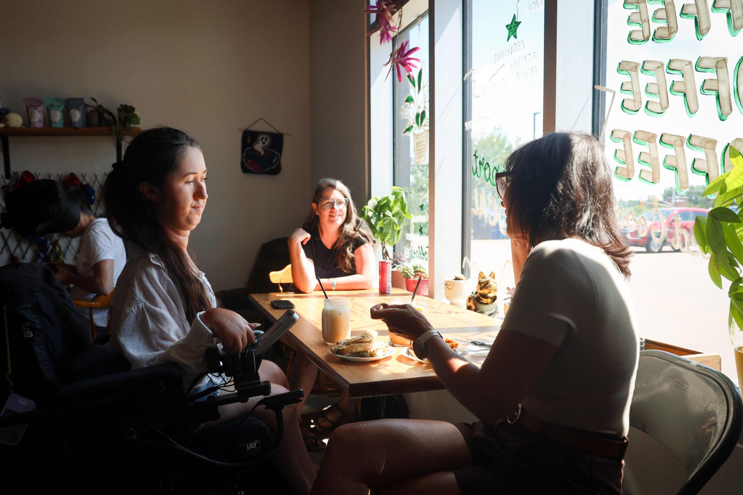 Tori Hunter, a wheelchair user and disabled travel blogger, sitting in her power wheelchair at a table inside the wheelchair accessible Ottawa Valley Coffee cafe in Petawawa, Ontario, chatting with friends over iced coffee and a panini.