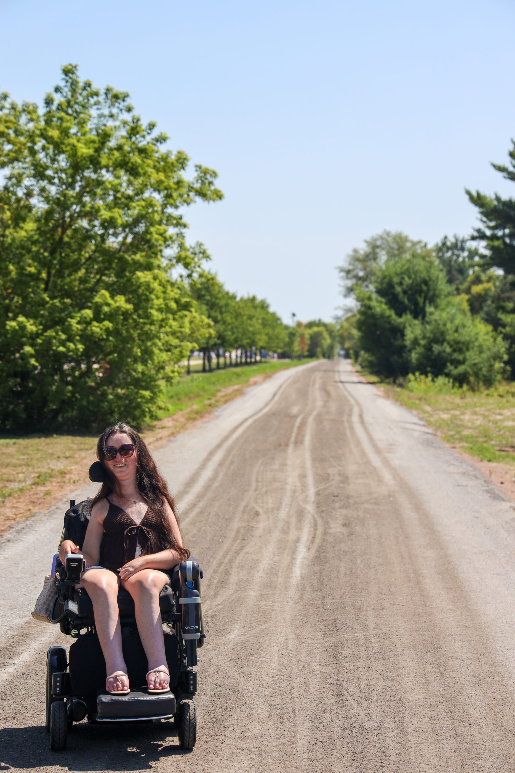 Tori Hunter, a wheelchair user and disabled travel blogger, riding her power wheelchair down a quiet tree-lined road in Petawawa, Ontario, on a sunny summer day.