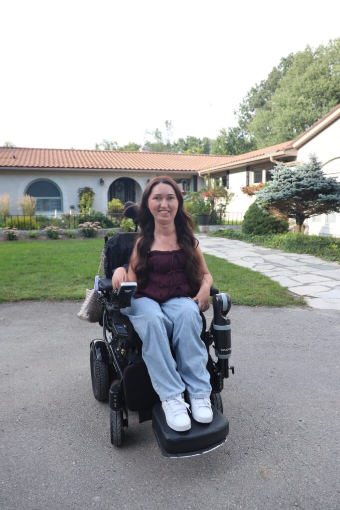 Tori Hunter, a wheelchair user and disabled travel blogger, smiling in her power wheelchair on the paved driveway in front of Country Oasis Accessible Retreat, a wheelchair accessible Airbnb in Innerkip, Oxford County, Ontario