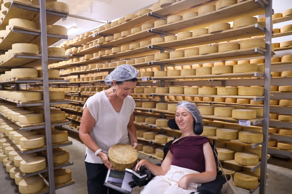 Tori Hunter, a wheelchair user and disabled travel blogger, in her power wheelchair as a cheesemaker shows her an aged wheel of cheese in the aging room during an accessible cheese factory tour at Gunn's Hill Artisan Cheese near Woodstock, Oxford County, Ontario