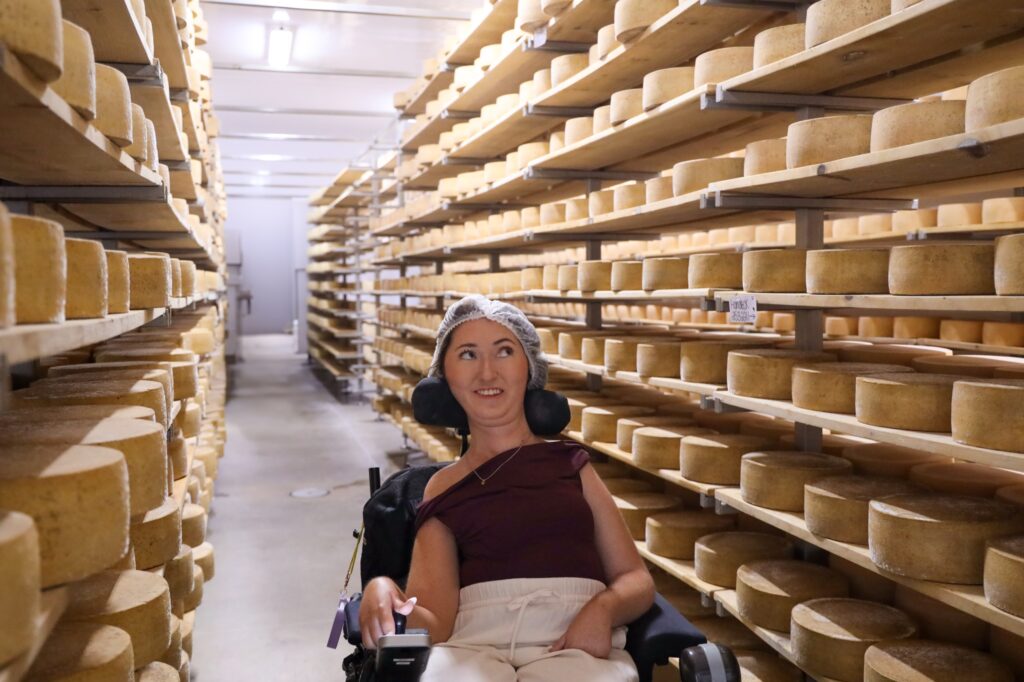 Tori Hunter, a wheelchair user and disabled travel blogger, smiling in her power wheelchair in a wide aisle between shelves of aging cheese wheels at Gunn's Hill Artisan Cheese near Woodstock, Oxford County, Ontario