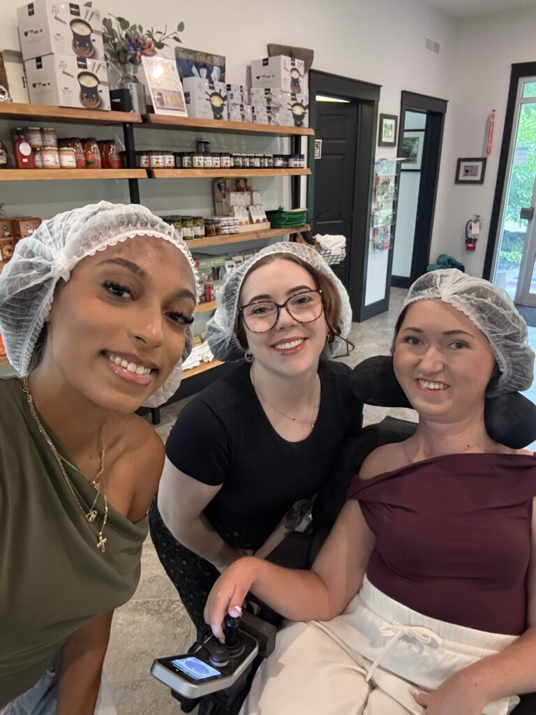 Tori Hunter, a wheelchair user and disabled travel blogger, taking a selfie with two friends in hairnets inside the shop at Gunn's Hill Artisan Cheese before an accessible cheese factory tour near Woodstock, Oxford County, Ontario