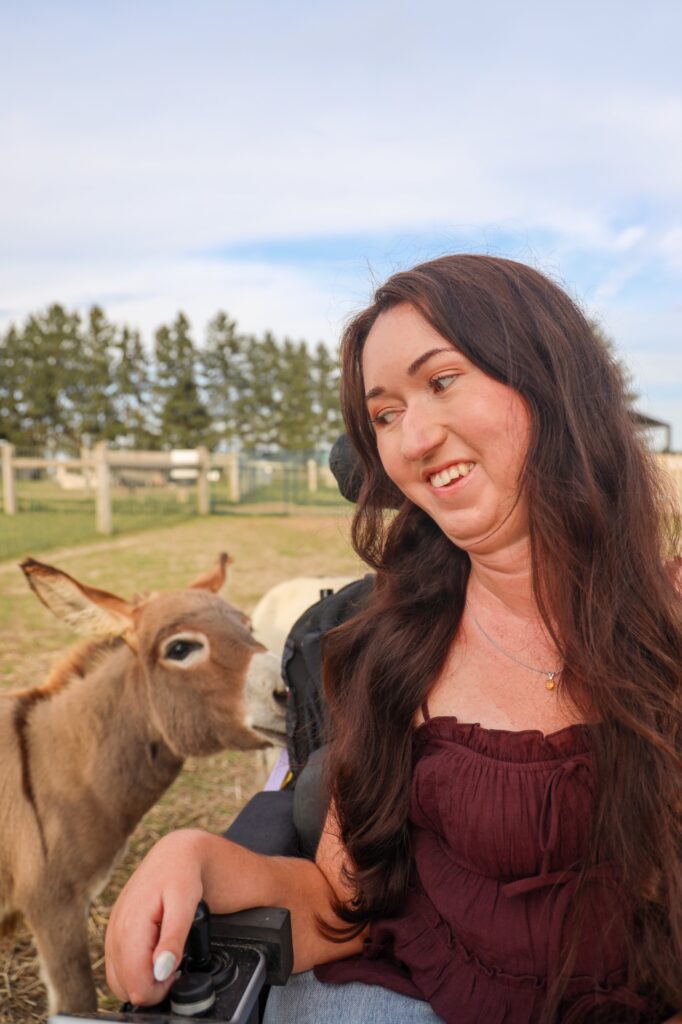 Tori Hunter, a wheelchair user and disabled travel blogger, smiling in her power wheelchair as a baby miniature donkey sniffs her chair at Udderly Ridiculous Farm Life in Oxford County, Ontario